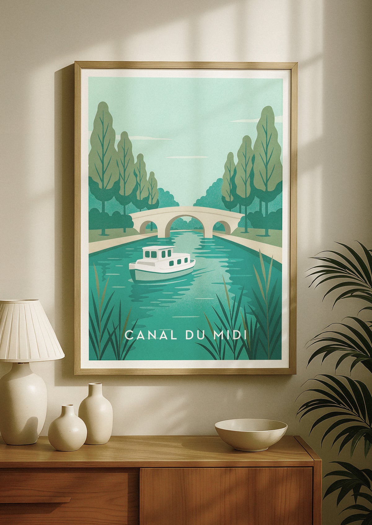 Ein gerahmtes Poster im Reiseposter-Stil mit dem Canal du Midi in Frankreich. Es zeigt ein Retro-Boot auf türkisfarbenem Wasser, Bäume und eine Steinbrücke. Das Poster hängt an einer beigen Wand über einem Holzkonsolentisch mit dekorativen Vasen und einer Lampe.