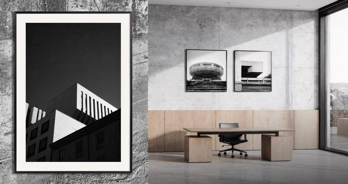 A black and white architectural art print in a white frame, displayed on a textured concrete wall next to a modern office interior featuring two matching framed prints above a desk.